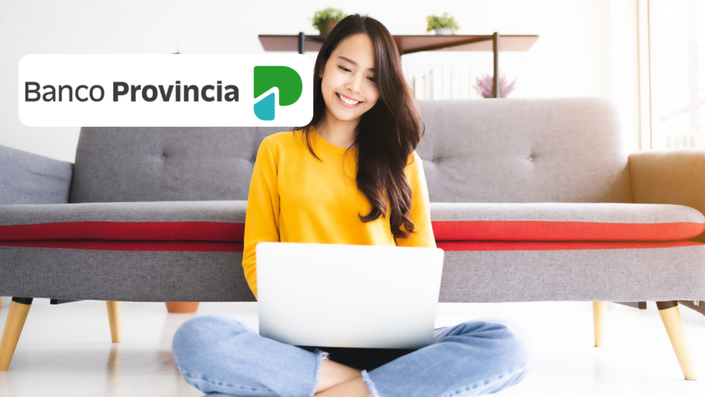 Mujer joven con cabello largo y oscuro, sonriendo mientras usa una laptop en el suelo de una sala bien iluminada. Lleva una blusa amarilla y está sentada con las piernas cruzadas frente a un sofá gris con detalles rojos.
