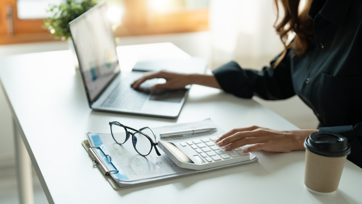 Mujer en su oficina con una libreta, gafas y una calculadora en su escritorio, mientras usa la computadora analizando tu presupuesto