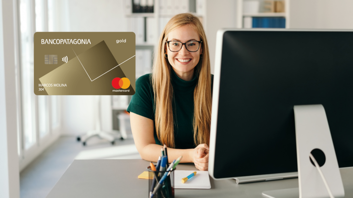 Mujer rubia con gafas, vistiendo un suéter negro, sonríe mientras trabaja en una computadora de escritorio en una oficina bien iluminada.