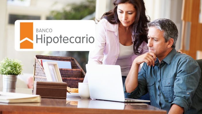 Una pareja madura revisa información en una laptop con expresiones de concentración. La mujer, de cabello oscuro y blusa rosada, está de pie mientras el hombre, de cabello canoso y camisa azul, está sentado.
