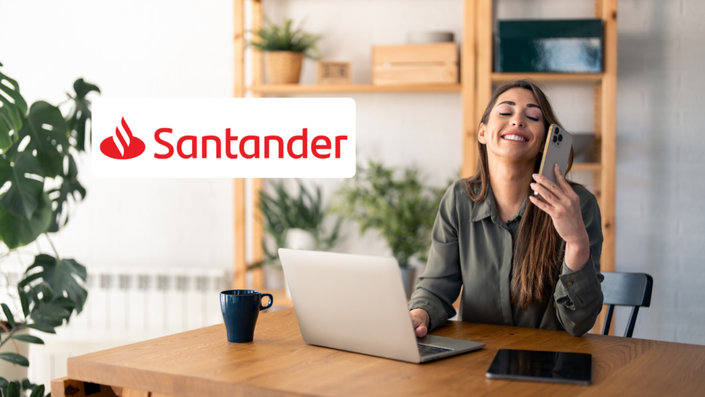 Mujer joven con camisa verde oliva sentada en una mesa de madera en un espacio de trabajo moderno y acogedor, sonriendo mientras sostiene un teléfono móvil. Frente a ella hay una laptop y una taza azul.
