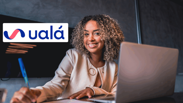 Mujer de negocios feliz trabajando en laptop en un ambiente de oficina, con el logo de Ualá visible, mostrando productividad y confianza.