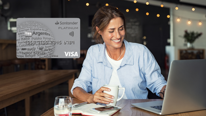 Mujer madura con camisa celeste sonriendo mientras trabaja en su laptop en un café iluminado con luces cálidas. La imagen destaca una tarjeta Santander Platinum Visa con diseño plateado y un globo terráqueo con nombres de países.