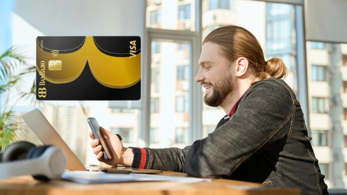 Hombre sonriente con el cabello recogido en un moño, usando una chaqueta oscura, sentado en una oficina luminosa mientras sostiene un teléfono móvil y trabaja en una laptop.