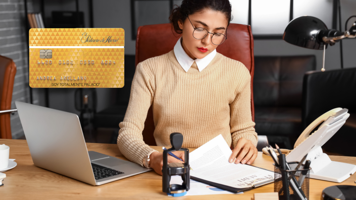 Mujer con gafas y suéter beige, concentrada mientras revisa documentos en un escritorio con una computadora portátil.