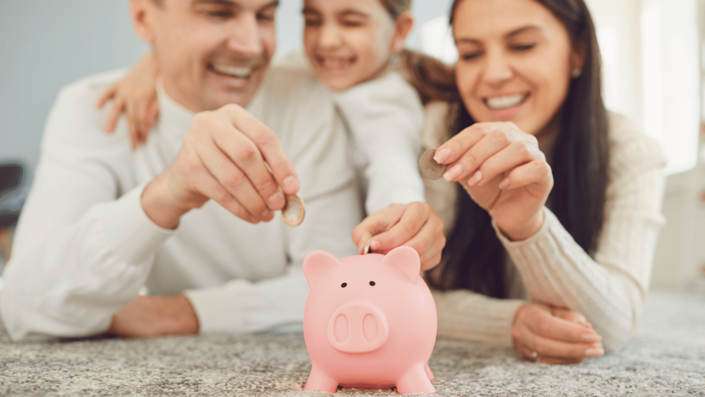 Familia sonriendo mientras deposita monedas en una alcancía rosada, simbolizando el ahorro en familia y la importancia de enseñar finanzas a los niños desde pequeños.