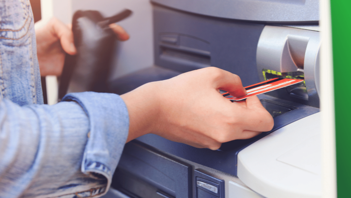 Persona insertando una tarjeta bancaria en un cajero automático. La persona viste una chaqueta de mezclilla y sostiene una billetera negra en la otra mano.