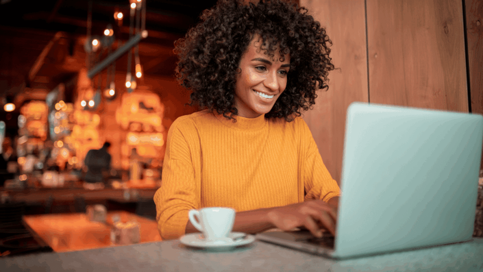Mujer joven usando una laptop en una cafetería, feliz mientras realiza compras en línea y busca ahorrar dinero desde México.