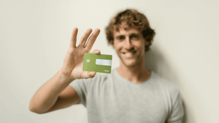 Un joven sonriente con remera gris clara sostiene una tarjeta de crédito color verde manzana en su mano derecha, mostrando cómo el acceso a productos financieros puede influir en tu Historial Crediticio. El fondo es de un color neutro y claro.
