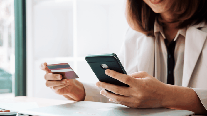 Una persona, vestida con una camisa clara y un saco de tono similar, está sentada frente a una laptop. Sostiene un celular con su mano izquierda y una tarjeta de crédito con la derecha, como si estuviera por realizar una compra online o gestionando sus finanzas, acciones que impactan directamente en su Historial Crediticio. Se la ve sonriendo levemente, sugiriendo una experiencia positiva.