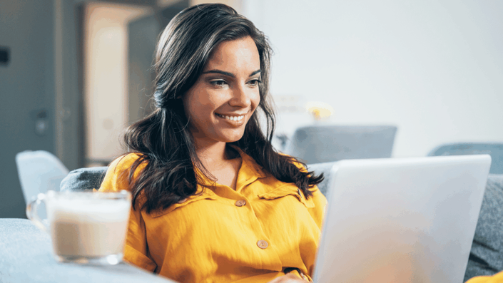 Mujer sonriente usando una laptop mientras disfruta de una bebida caliente, mostrando una experiencia positiva al gestionar su préstamo personal en línea desde la comodidad de su hogar.