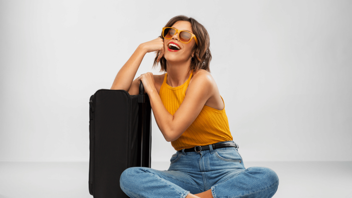 Mujer sonriente, con gafas de sol, sentada junto a una maleta, con una tarjeta de crédito para viajeros en la mano, preparándose para su próximo viaje.